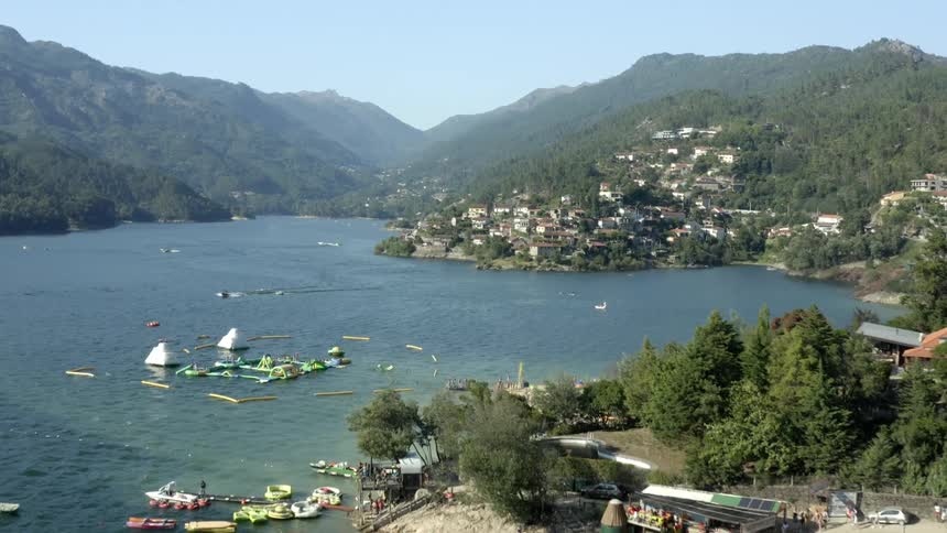 People around beach as others ride boats leaving wakes near town sitting on slope near shore among forest with other mountains in background