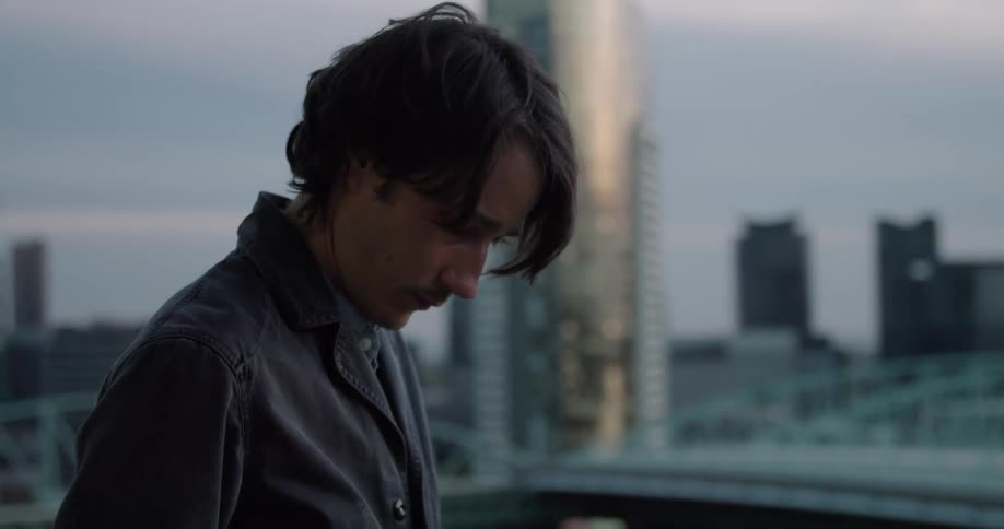 A man stands on a rooftop balcony texting on a phone while looking at the city skyline