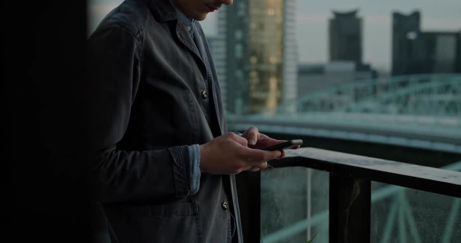 A man in a balcony using a smart phone with buildings the sky and clouds in the background