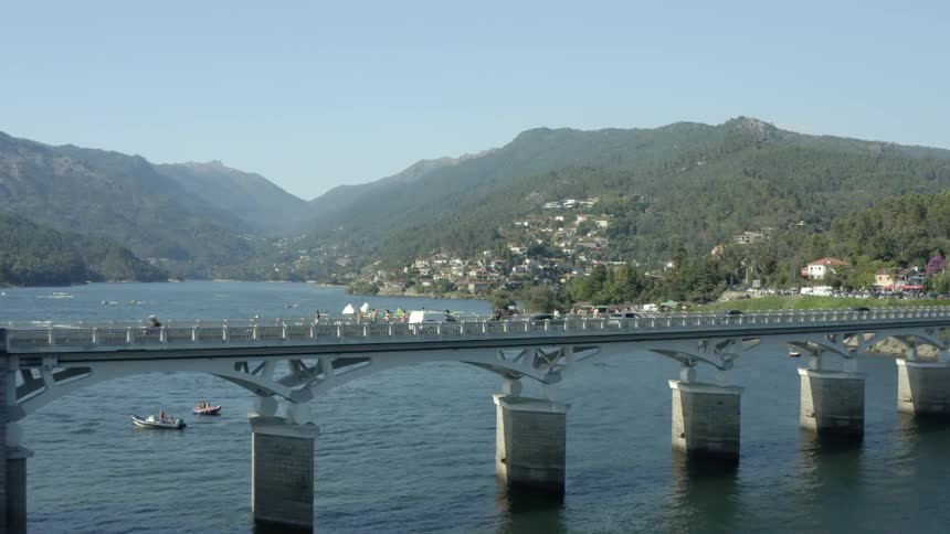 People and cars traveling across a bridge over a river near a town on a mountain side