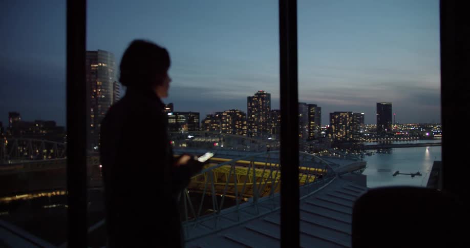 Man using smartphone in dim lit office is silhouetted against the sky next to stadium at dusk