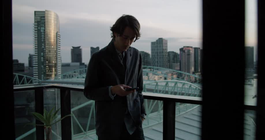 A man is texting on his phone as he stands on a balcony in a city