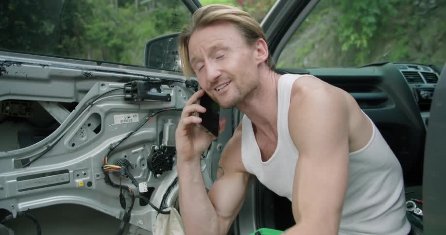 A man is sitting on the edge of his car seat and talking on the phone