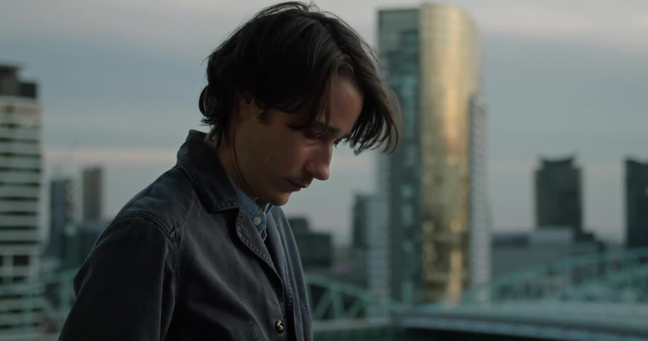 A man is standing on a balcony looking at tall city buildings and texting on his cell phone