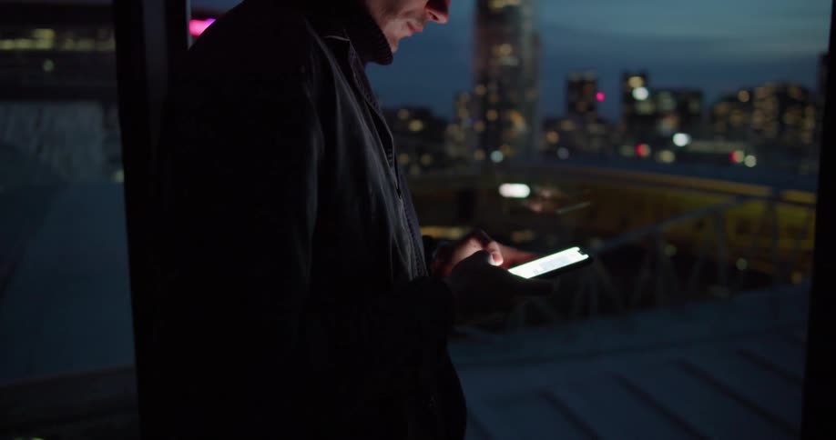 Man stands near window looking out onto city at night while texting and smiling