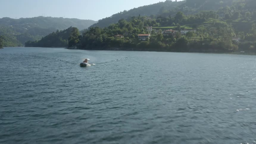 People ride boat leaving wake over water away from red roofed villa and mist covered mountain slopes