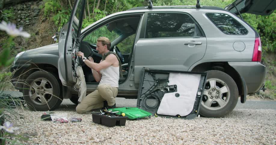 Man crouches working on interior of door of car parked by road on pebbles