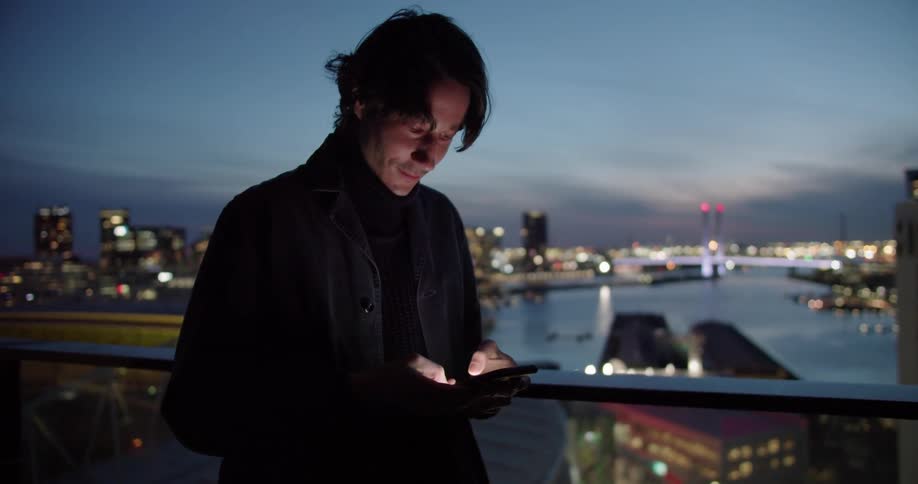 A man is standing on a balcony texting on his phone with a big city in the distance