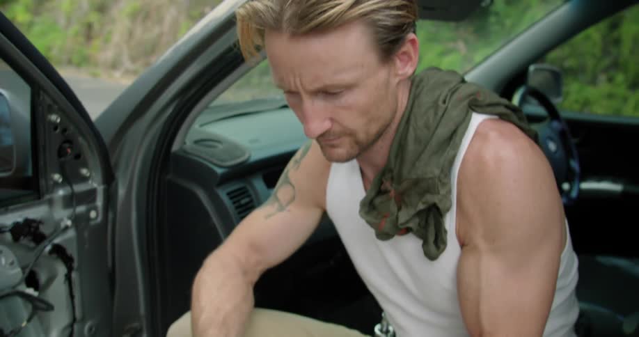 Muscular blonde man sitting on a car parked at the side of the road texting on the phone