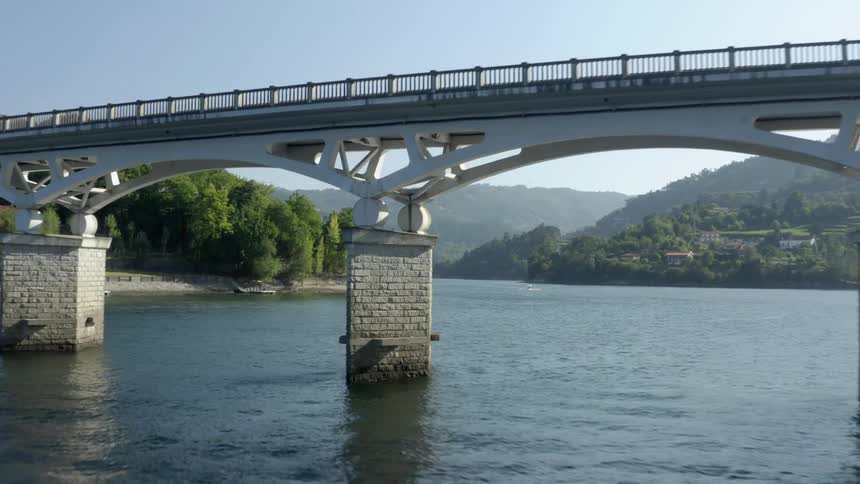 An aerial view of flying under an arch bridge over a river with homes along the riverbank and mountainside in the daytime