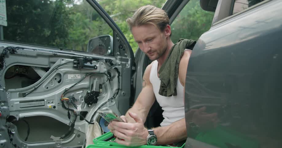 A man sitting in a broken down car in front of a forest uses his cell phone to call someone