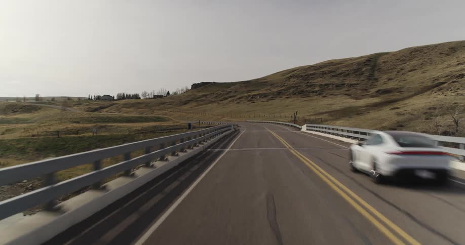 electric sports car drives along highway over bridge passing hill toward buildings and trees on top of hill
