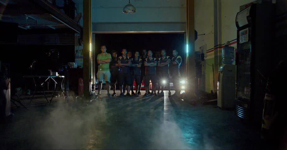 A race team stands posed in a garage doorway with arms crossed and headlights shining behind them
