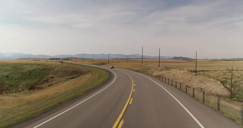 Electric Car drives along highway past fenced pastures paralleling power lines while heading to mountains on foggy horizon