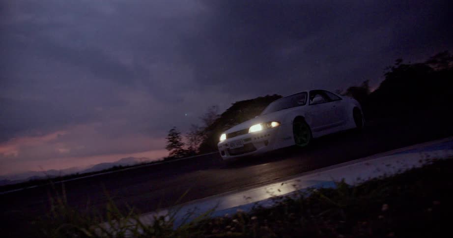 A white car is driving on a road at night with the headlights on and trees in the distance
