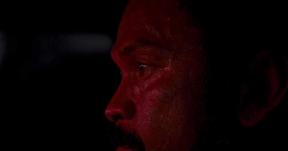 A flashing red and blue light highlights a mans skin texture as he sits in a dark room