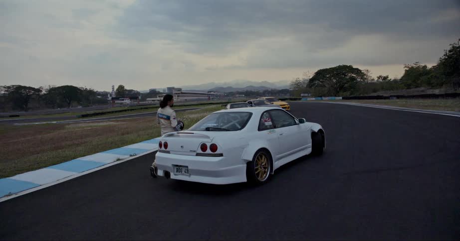 A race driver walks towards the driver side door of a car parked on a race track while carrying a helmet