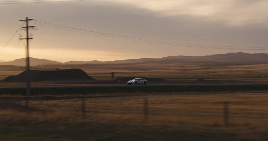 Electric car speeding along a side road and turns onto the main highway without stopping in a desolate landscape with the sun setting behind mountains in the background