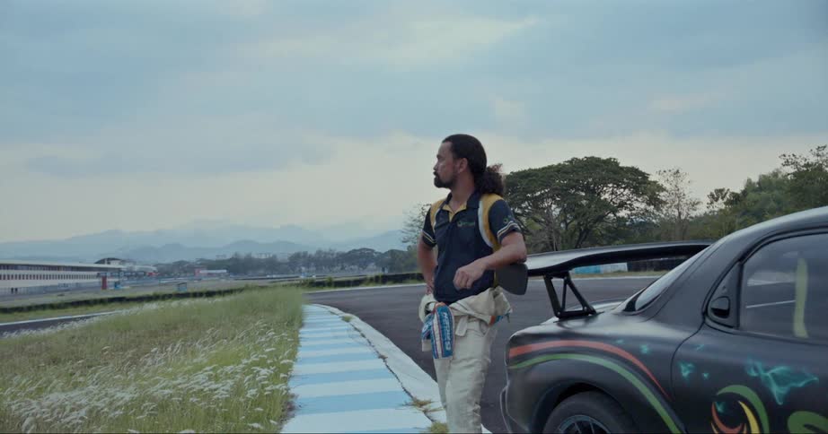 A man with coveralls rolled down to his waist stands next to a race car leaning on its spoiler looking at the track