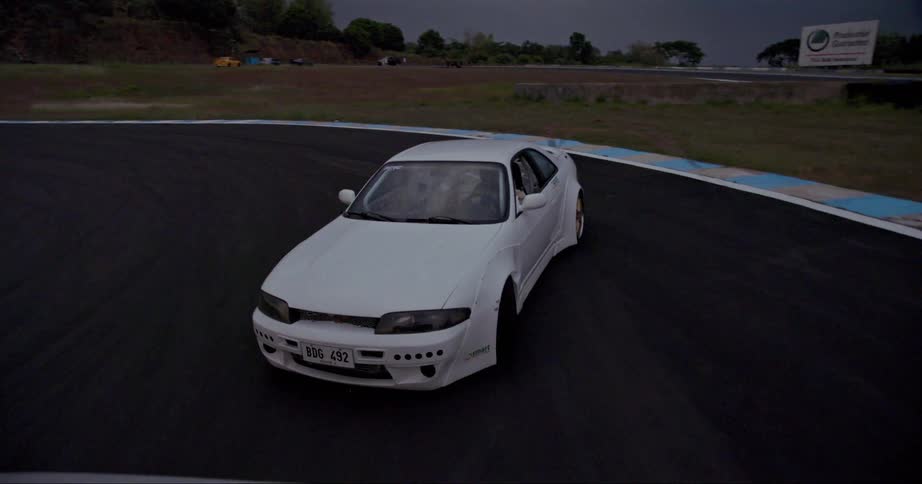 White car follows truck around track with hill and billboard in background