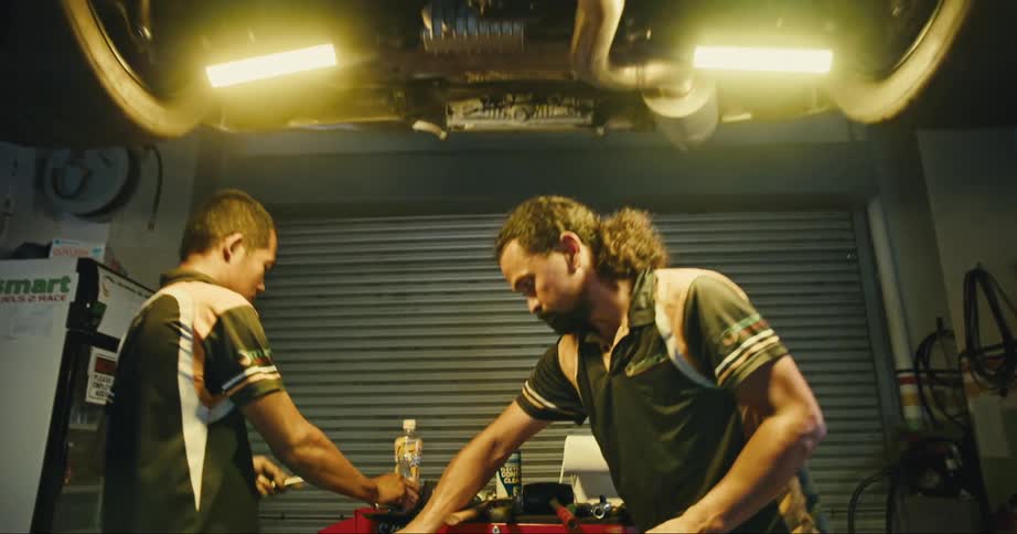 Two men work on a car up on a lift in a garage with lights glowing from the undercarriage