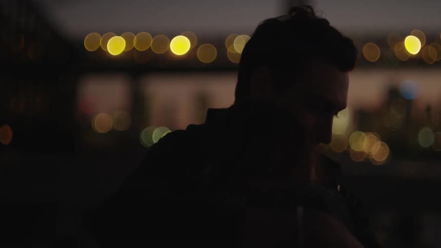 A man is standing in the dark near a lit bridge