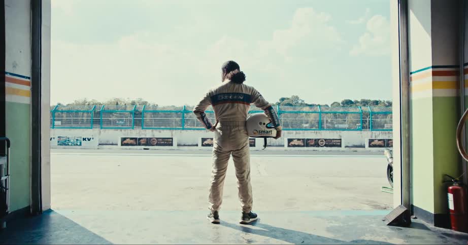 A race car driver holds his helmet under his arm while standing in the entrance to a garage at a race track