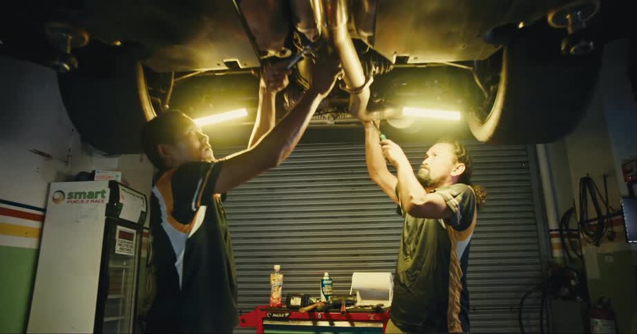 A man strains as he tightens a clamp underneath a car on a lift in a garage bay