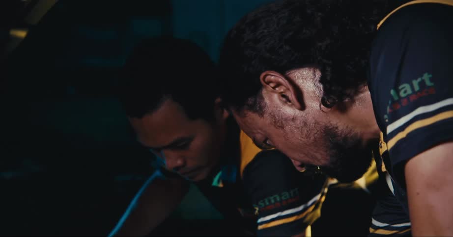 Two men with matching jerseys in a garage working on a vehicle with a wall in the background