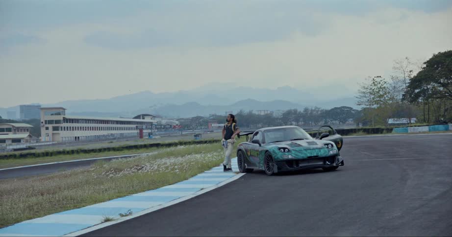 Man stands leaning on parked sports car with fenced building and foggy mountains in background