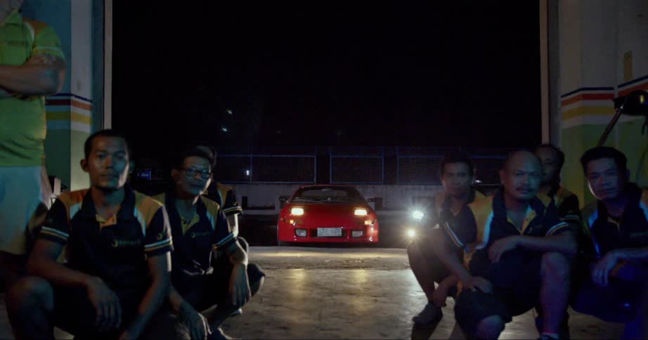 Sports car headlights shine into a garage bay where a mechanic team is posed