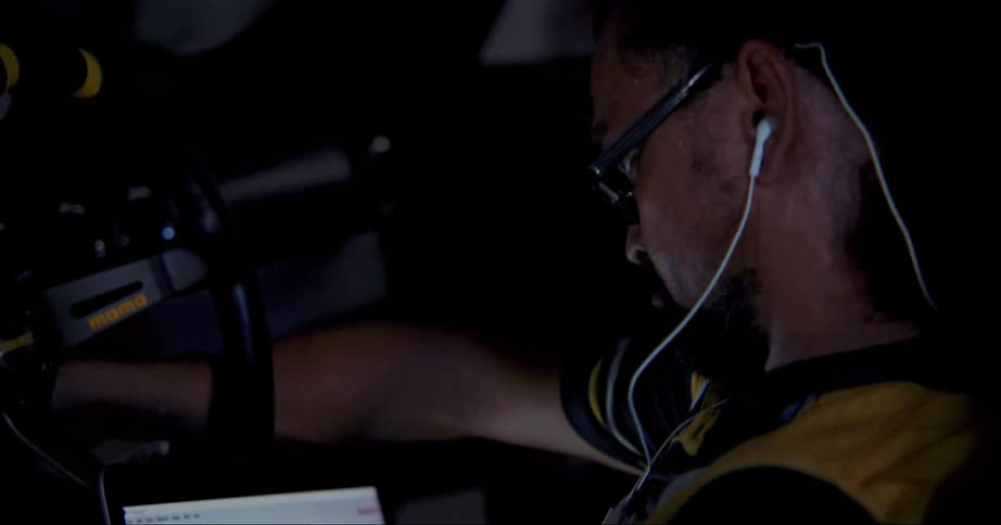 A man with glasses inside a vehicle with a steering wheel working on it what a laptop on his lap
