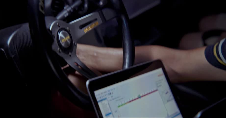 A man with glasses inside a vehicle with a steering wheel working on the vehicle what a laptop on his lap