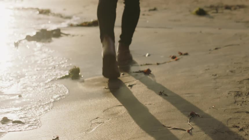 Person wearing laced ankle boots walks along beach past kelp and surf