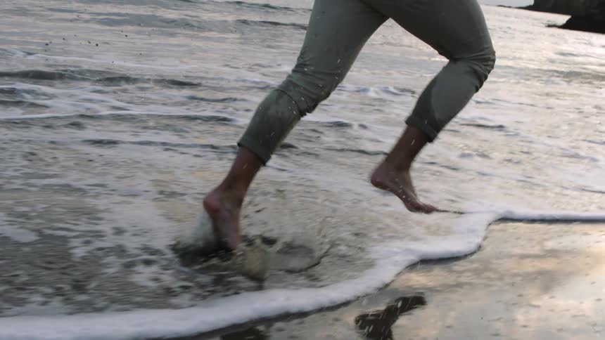 A man is jogging barefoot on a beach while wearing pants and a shirt during the evening
