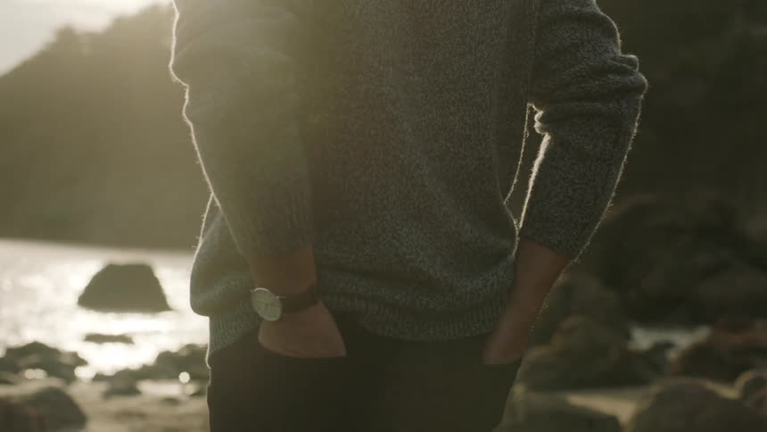 Man with gray sweater and black pants puts his hands in his back pockets walking on the coast next to sea