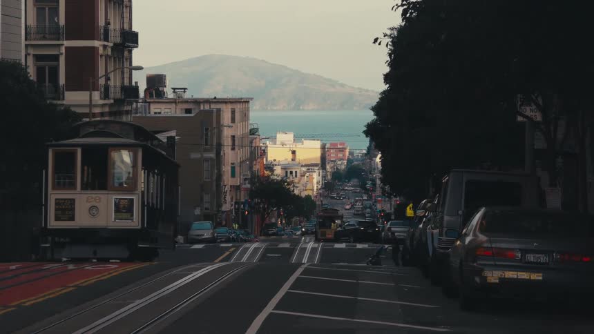 Pedestrian pushes baby carriage across hilly street as streetcars and other traffic moves along intersection and road with ocean in background