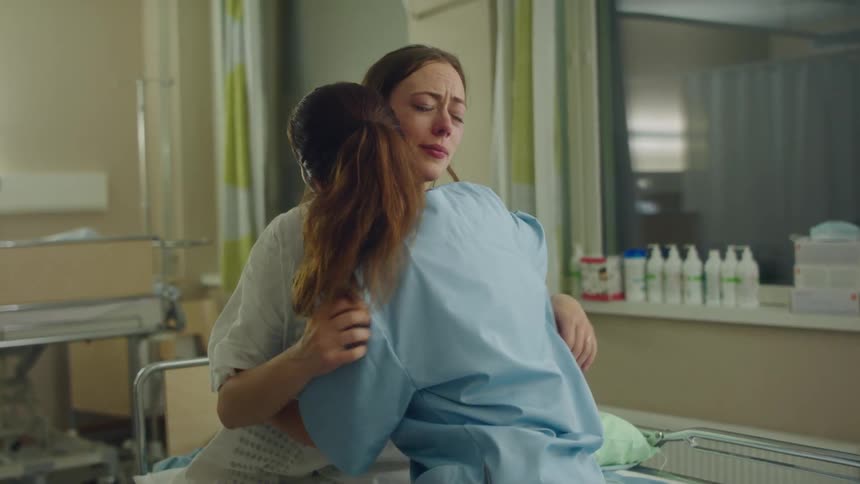 Female nurse with pony tail and scrubs cuddles female patient wearing white post surgery shirt and crying in hospital room