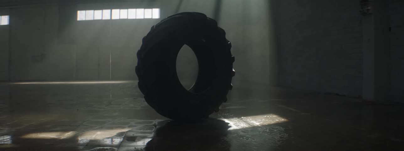 A large truck tire is standing by itself on a garage floor as light from the windows shines on it