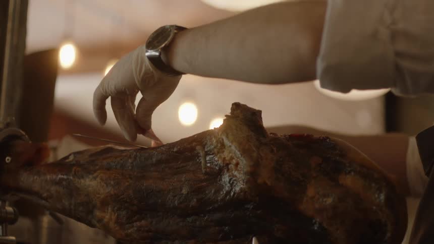 A man carves paper thin slices of ham from a pork leg