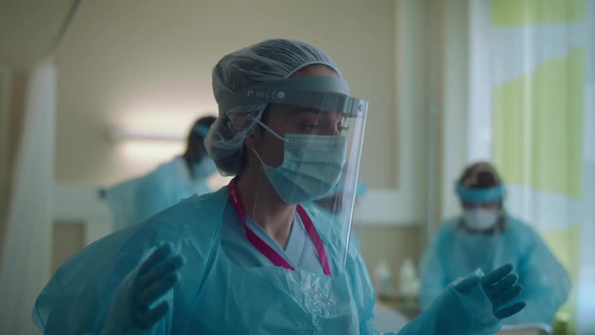 Nurse stands unmoving in scrubs and protective gear as other medical personnel move around hospital bed in background
