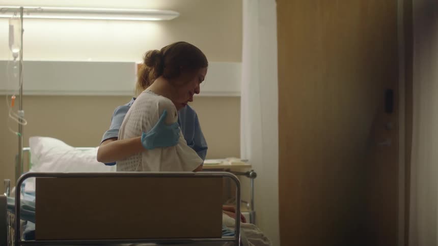 Female nurse with scrubs sympathizes with woman sitting on bed crying in hospital room