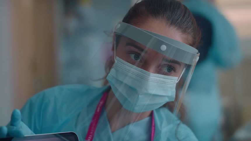 A nurse with a mask face shield scrubs and gloves in a hospital room with a tablet