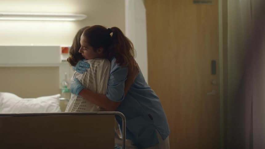 Patient sitting on side of hospital bed cries and nurse hugs her to provide comfort