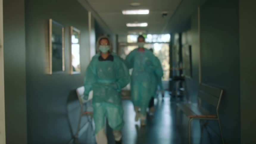 Five doctors with masks walking down a hospital hallway with benches with a door in the background