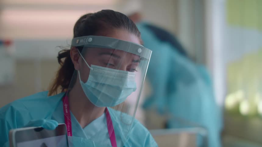 A nurse with a mask face shield scrubs and gloves in a hospital room with a tablet