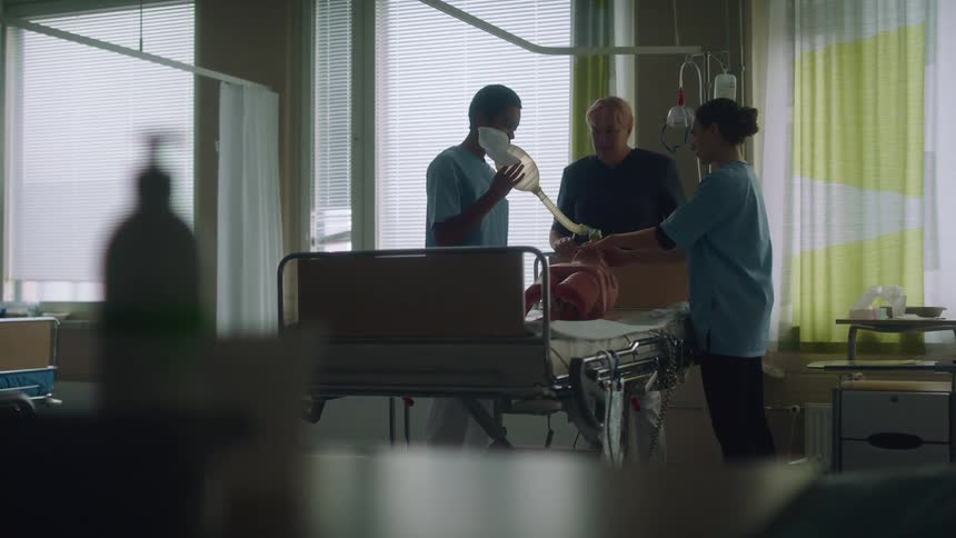 Doctor with short hair blue shirt demonstrates nurses how putting mask on mannikin