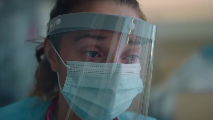 A nurse wearing a face mask and shield is staring worriedly at a patient while in a hospital