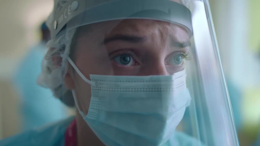 A woman wearing scrubs mask and face shield looks nervous and worried