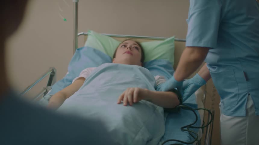 A woman in a hospital room laying in a hospital bed as a nurse with scrubs and gloves gets ready to check her blood pressure
